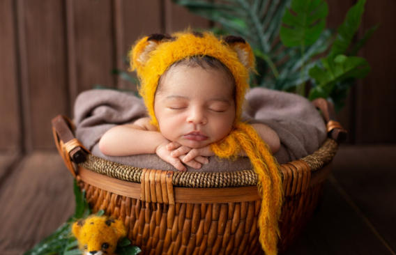 infant sleeping pretty baby boy yellow animal shaped hat inside brown basket along with green leafs wooden room